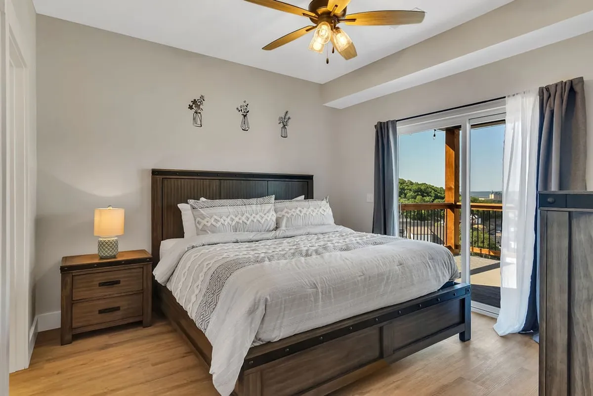 A serene bedroom with a large bed, soft white linens, a nightstand topped with decorative flowers, and a sliding glass door that opens to a private deck. Natural light filters into the room, giving it a peaceful, inviting atmosphere.