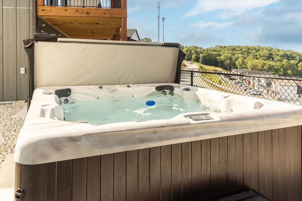 A multi-level exterior deck with a private jacuzzi set into the decking, surrounded by scenic Ozark landscapes. There is ample outdoor seating, inviting guests to relax and enjoy views or socialize, with the jacuzzi as a focal point. Subtle lighting and the tranquil atmosphere make it ideal for day or night relaxation.