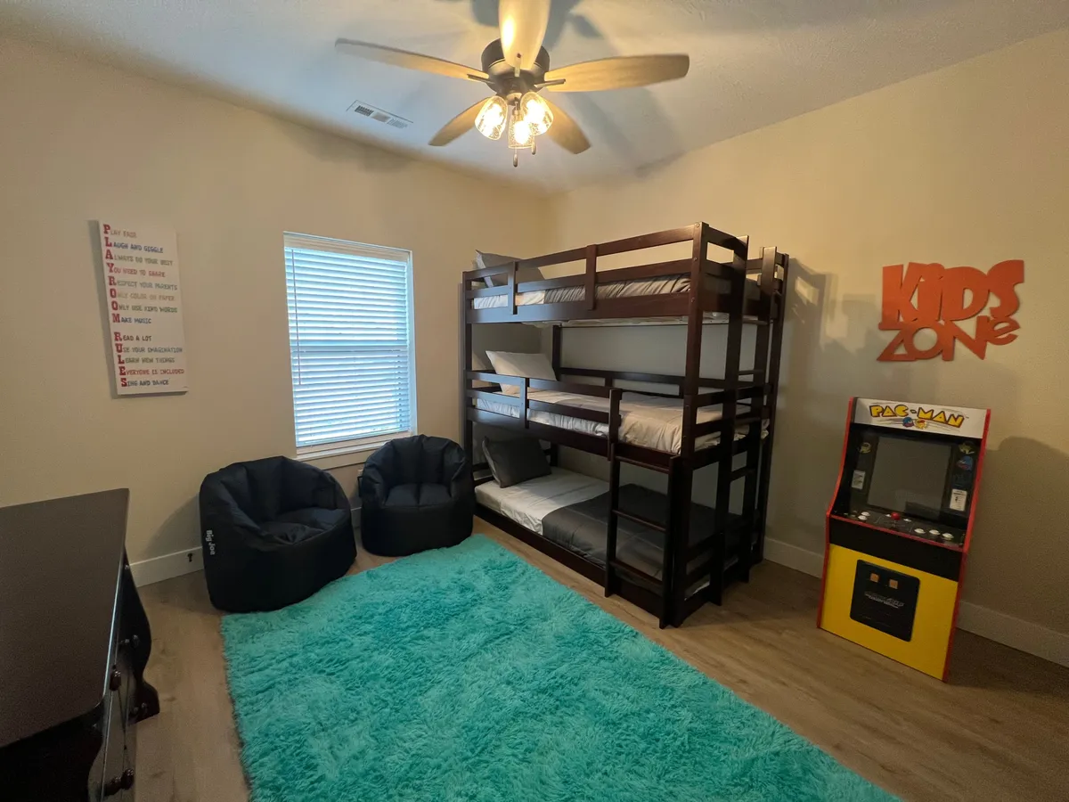 A lively and colorful kids’ bunk room featuring triple bunk beds, bean bags, an arcade game machine, and a playful 'kids zone' sign. The space is designed for fun sleepovers and engaging activities, making it inviting for children.