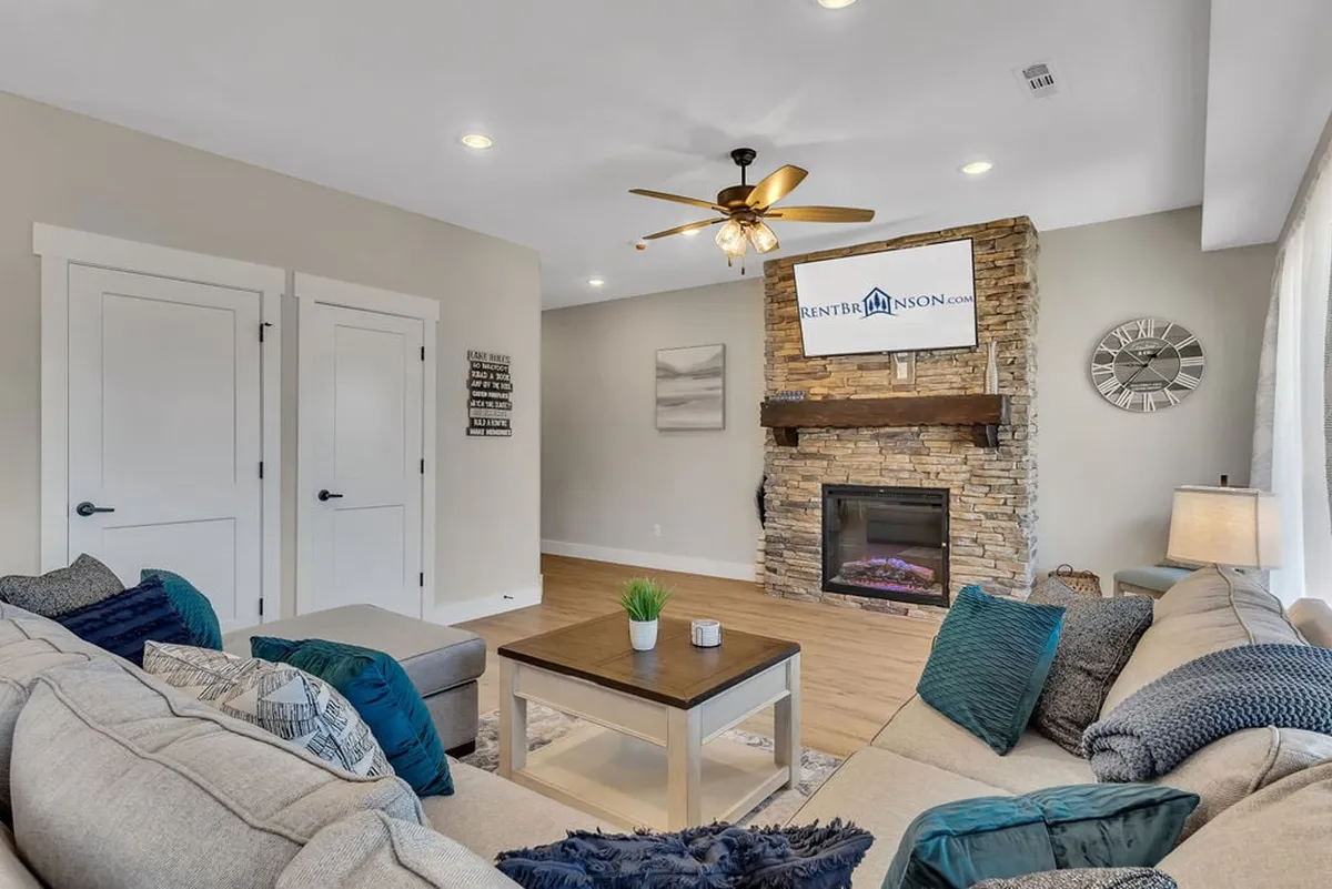 This cozy living room features a comfortable sectional sofa, a striking stone fireplace, a coffee table, a wall-mounted television, and large windows that let in plenty of natural light. The room is designed for relaxing with family, enjoying movie nights, or gathering for intimate conversations.