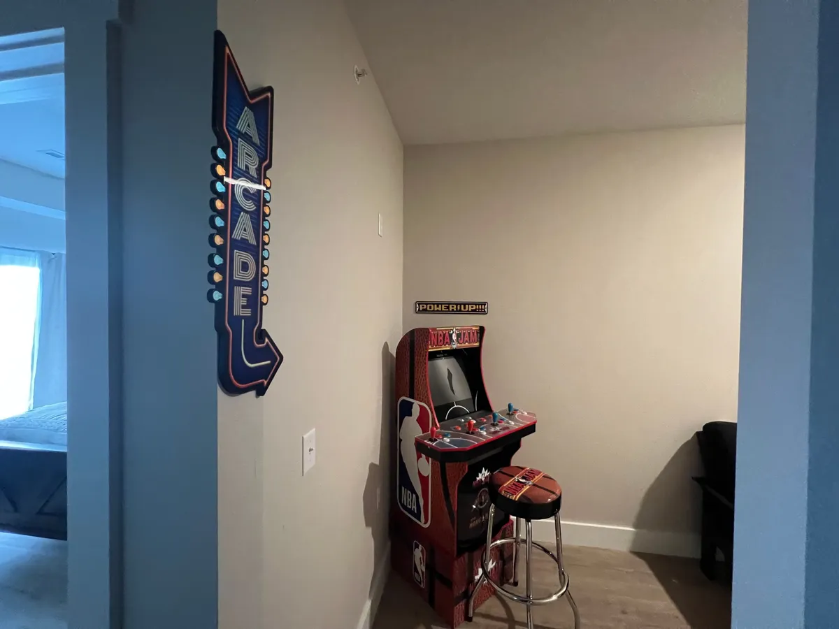 An energetic interior view of a game room featuring an arcade sign, an NBA Jam arcade machine, and a bar stool. The space is designed for fun, friendly competition, and nostalgic gaming experiences with family or friends.