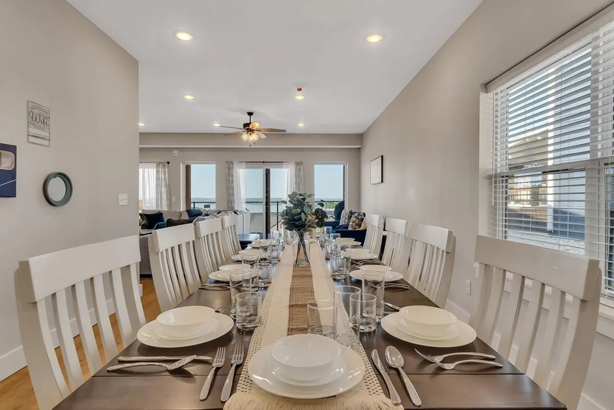 The image showcases a long dining table elegantly set for a meal, bathed in natural light streaming through expansive windows. Decorative plants add a touch of warmth and freshness, creating an inviting space perfect for group gatherings.