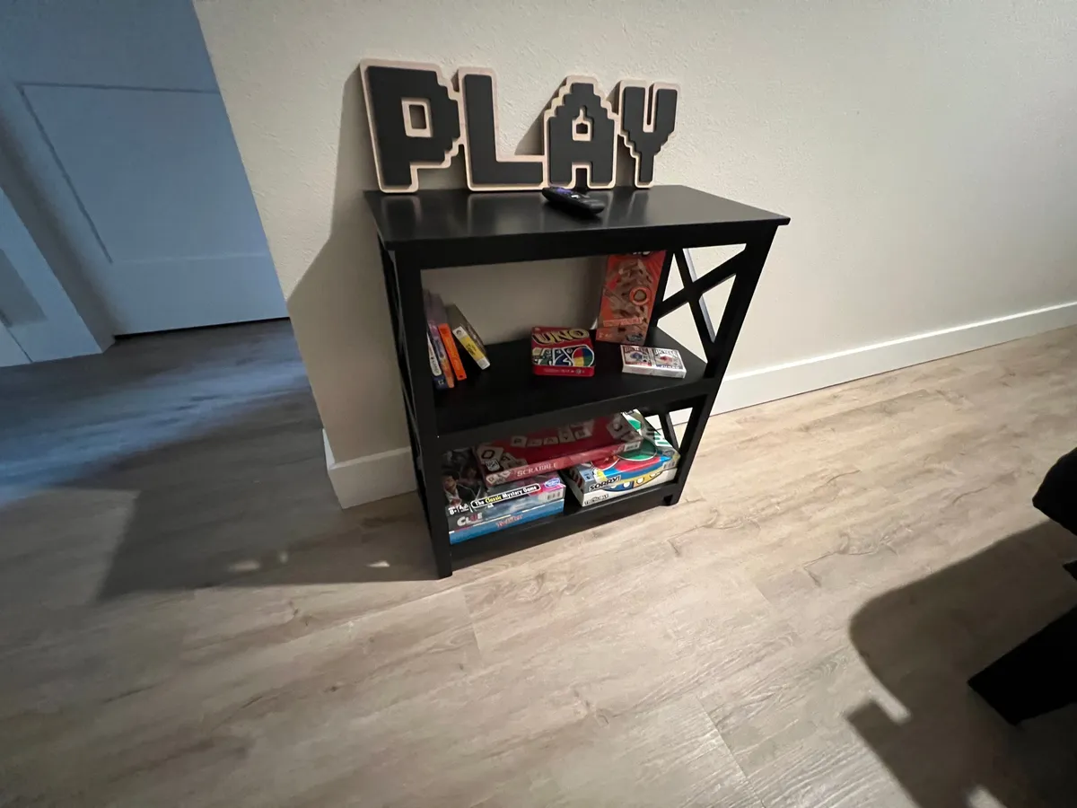 A welcoming corner of the game room featuring a black shelf filled with board games, a decorative 'PLAY' sign above, and a setting arranged for group games and family gatherings.
