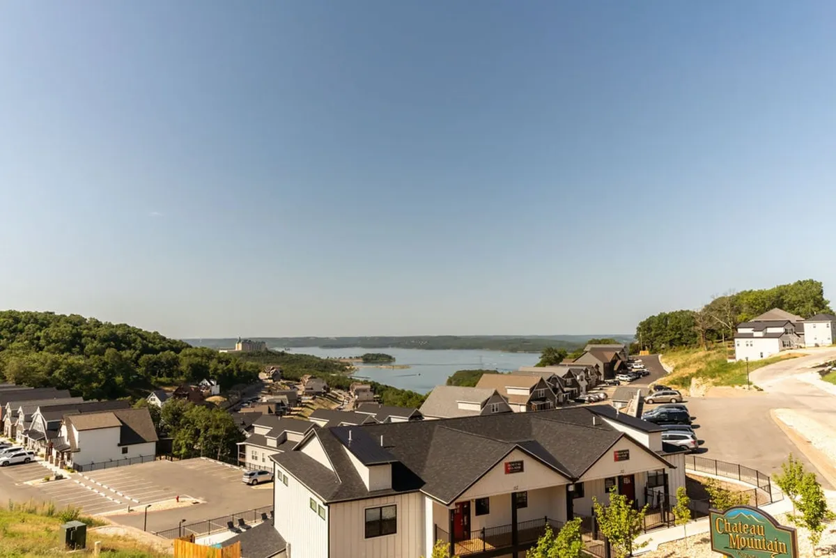 A sweeping view of Table Rock Lake with the serene waters, surrounding green hills, and nearby residential buildings. The image captures the lake as a focal point, with clear skies and gentle sunlight suggesting a relaxed vacation environment.