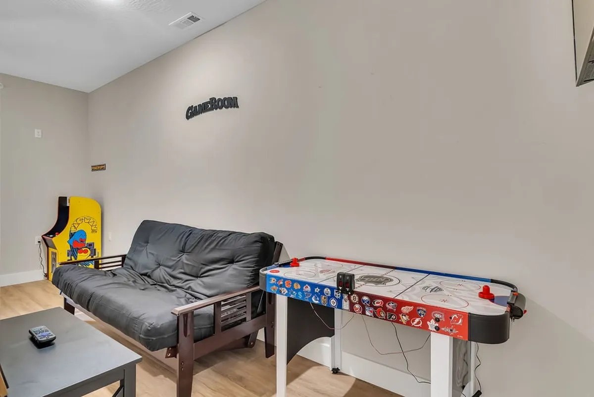A bright and spacious game room with a futon couch, a full-sized air hockey table, and a classic arcade game machine. The space is designed for both active gaming and lounging, with ample space for friends and family to gather.