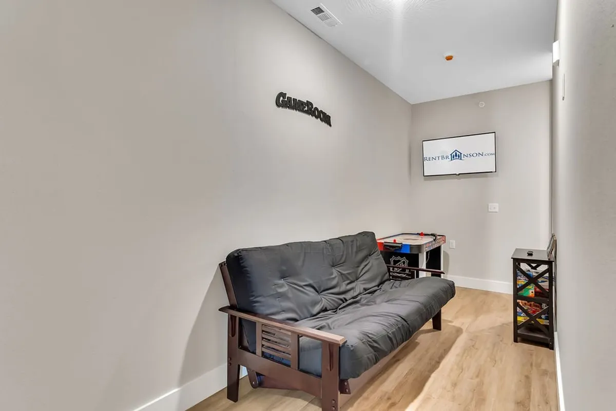 A relaxed and inviting game room scene with a futon sofa, a wall-mounted 'game room' sign, a television, and a table set up for board or video games. The atmosphere is cozy, ideal for both screen-based gaming and group play.